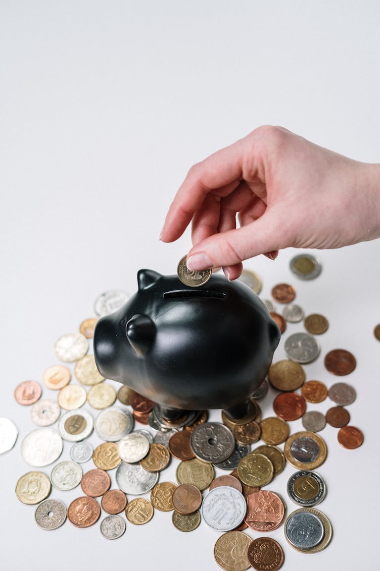 Hand inserting coin into black piggy bank among scattered coins, symbolizing savings and financial planning.