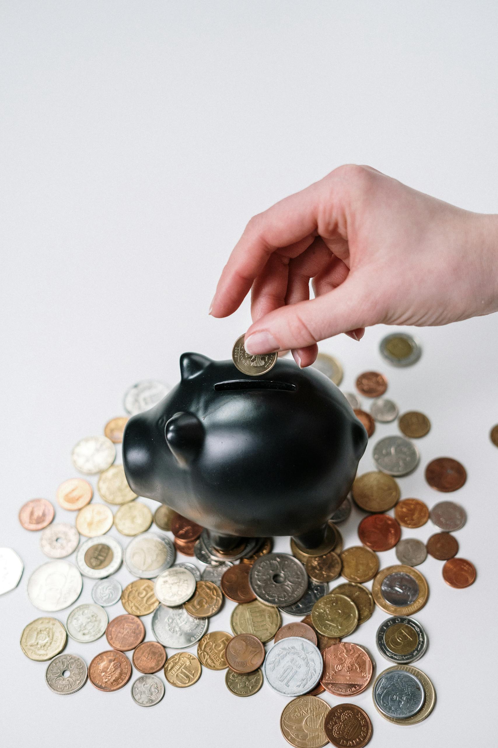 Hand inserting coin into black piggy bank among scattered coins, symbolizing savings and financial planning.